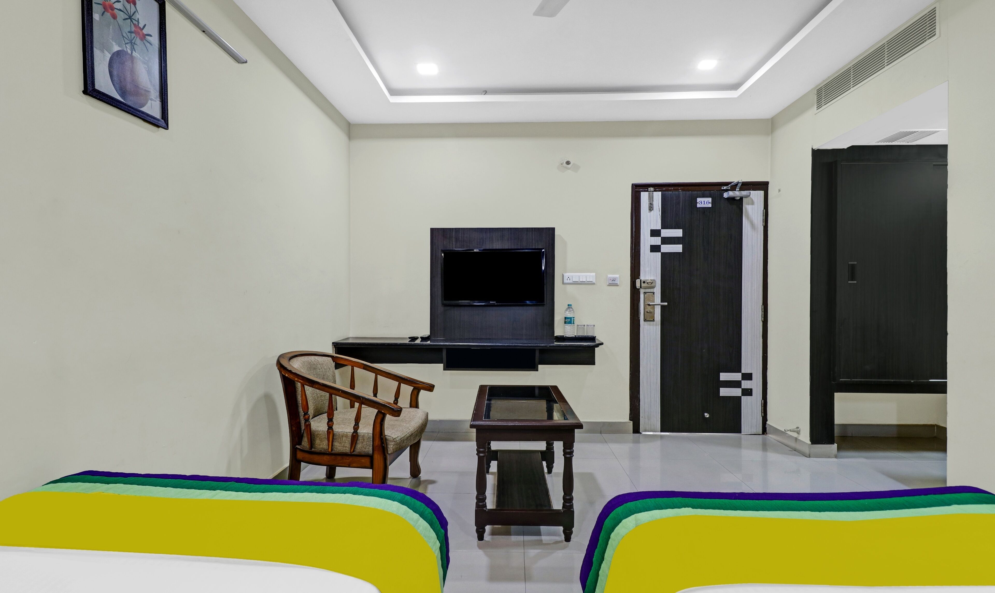A hotel room with a flat-screen TV mounted on the wall, a wooden chair, a small table, and a bed with colorful bedding.
