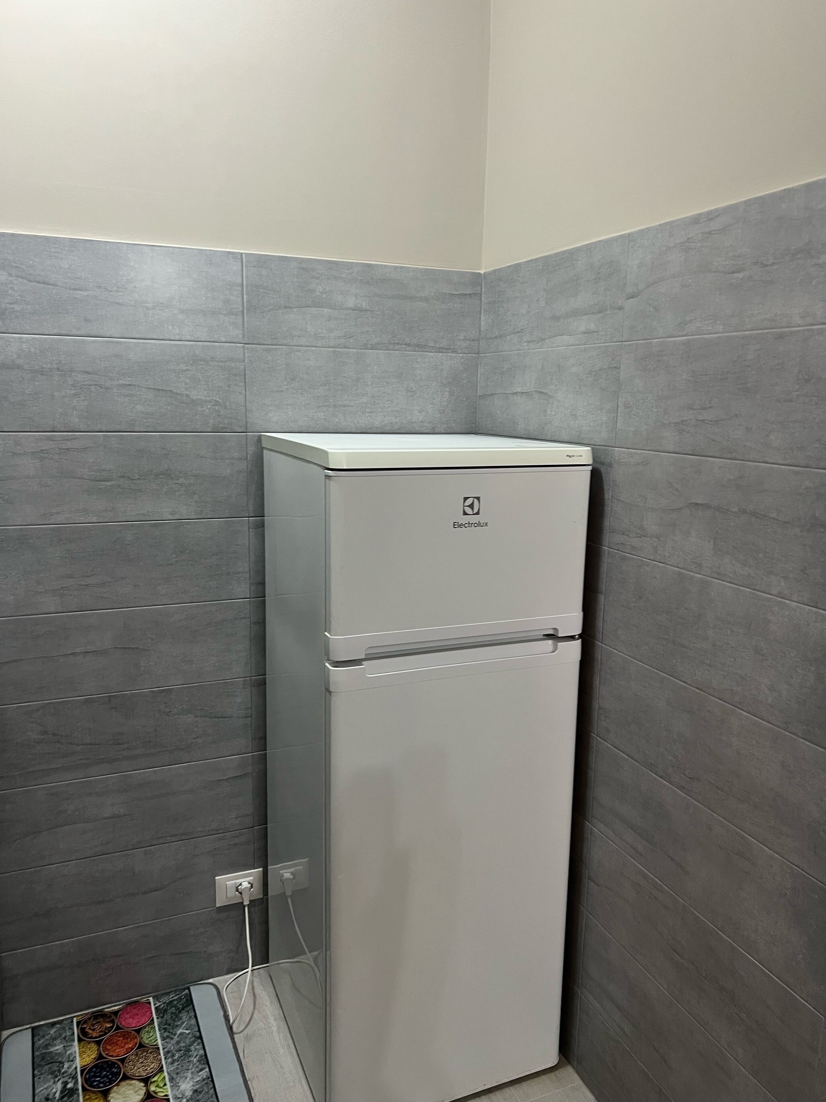 A compact, white refrigerator with a brand logo, situated in a corner of a room with grey tiled walls.