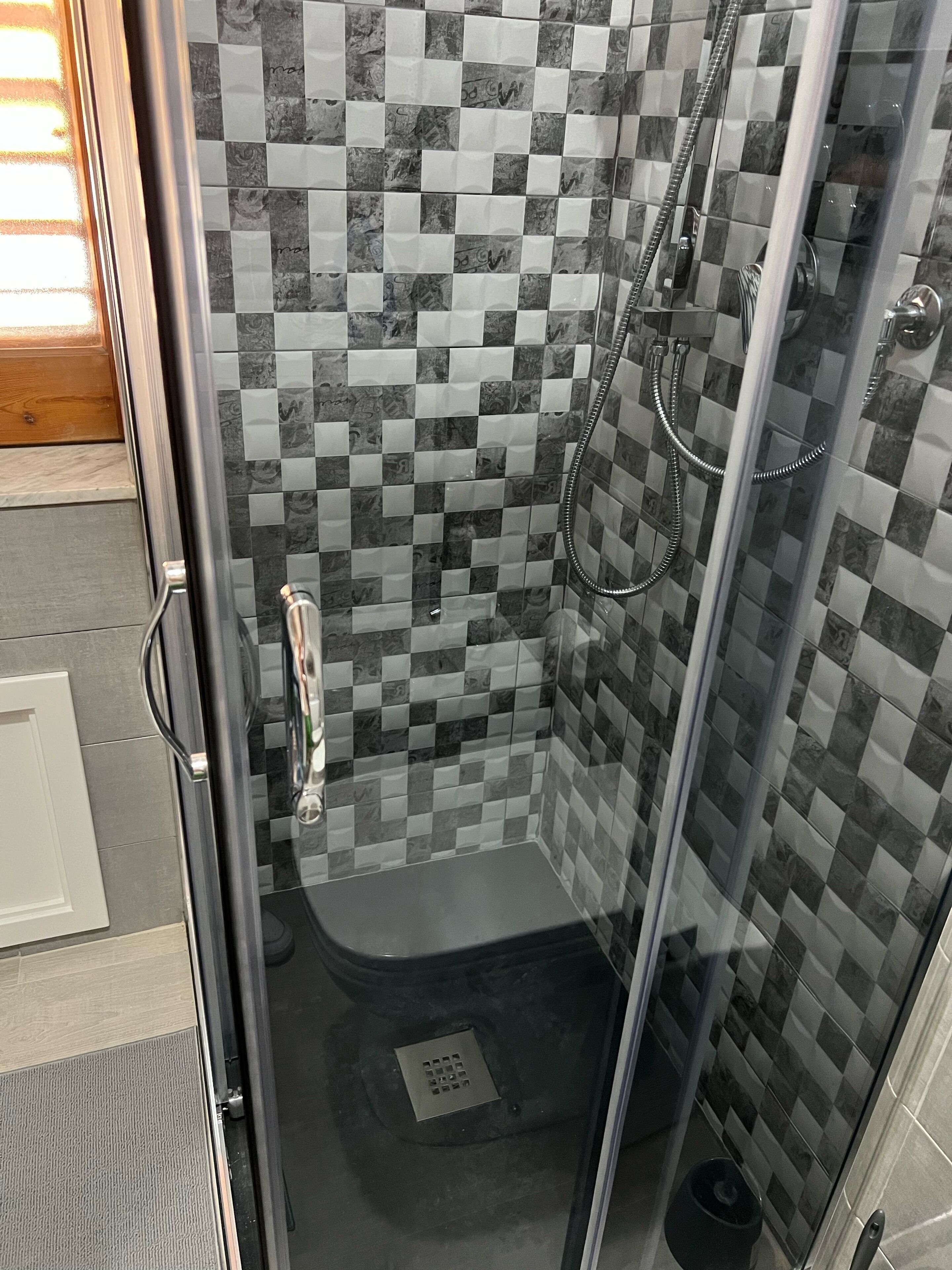 Una ducha con cerramiento de vidrio, paredes de azulejos a cuadros y desagüe visible.