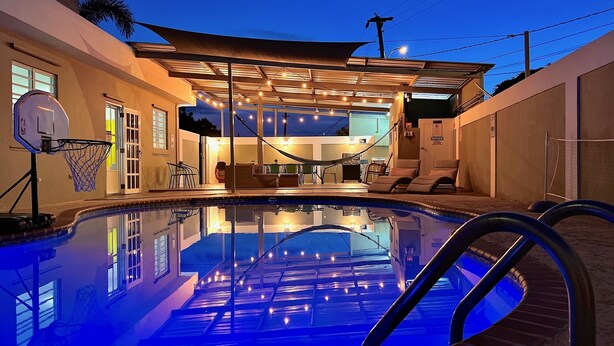 Piscina com aro de basquete, cadeiras de descanso e área de estar coberta com luzes de corda.