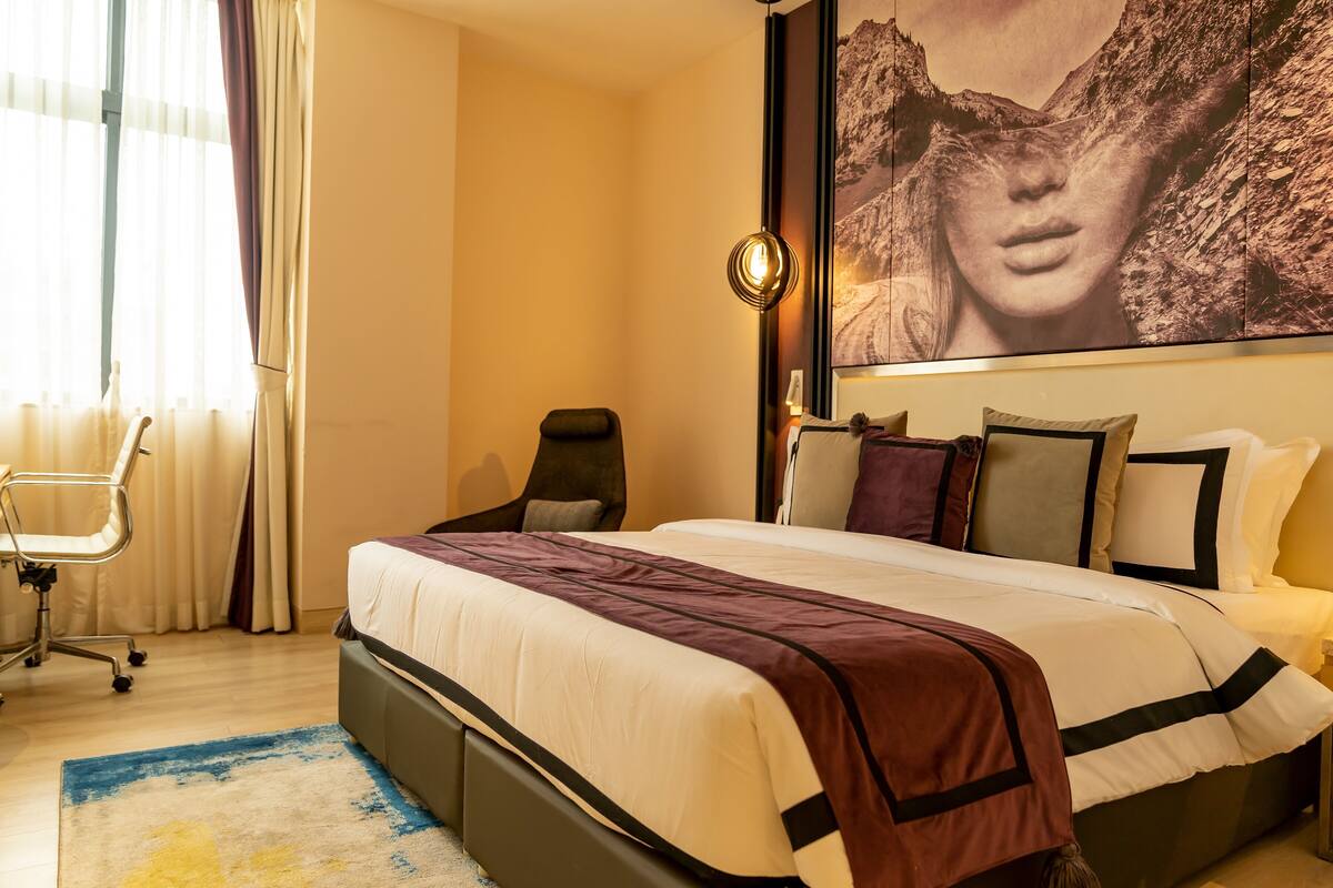 A hotel room with a bed, a chair, a desk, and a large painting on the wall.