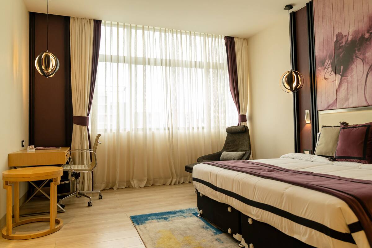 A hotel room with a bed, a desk, a chair, and a large window with curtains.