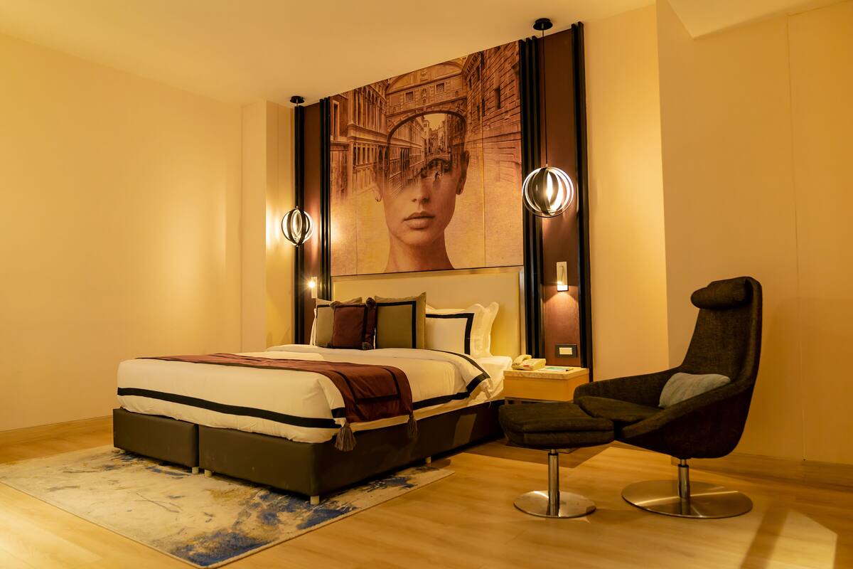 A modern bedroom with a large bed, bedside lamps, a chair, and a large wall art piece.