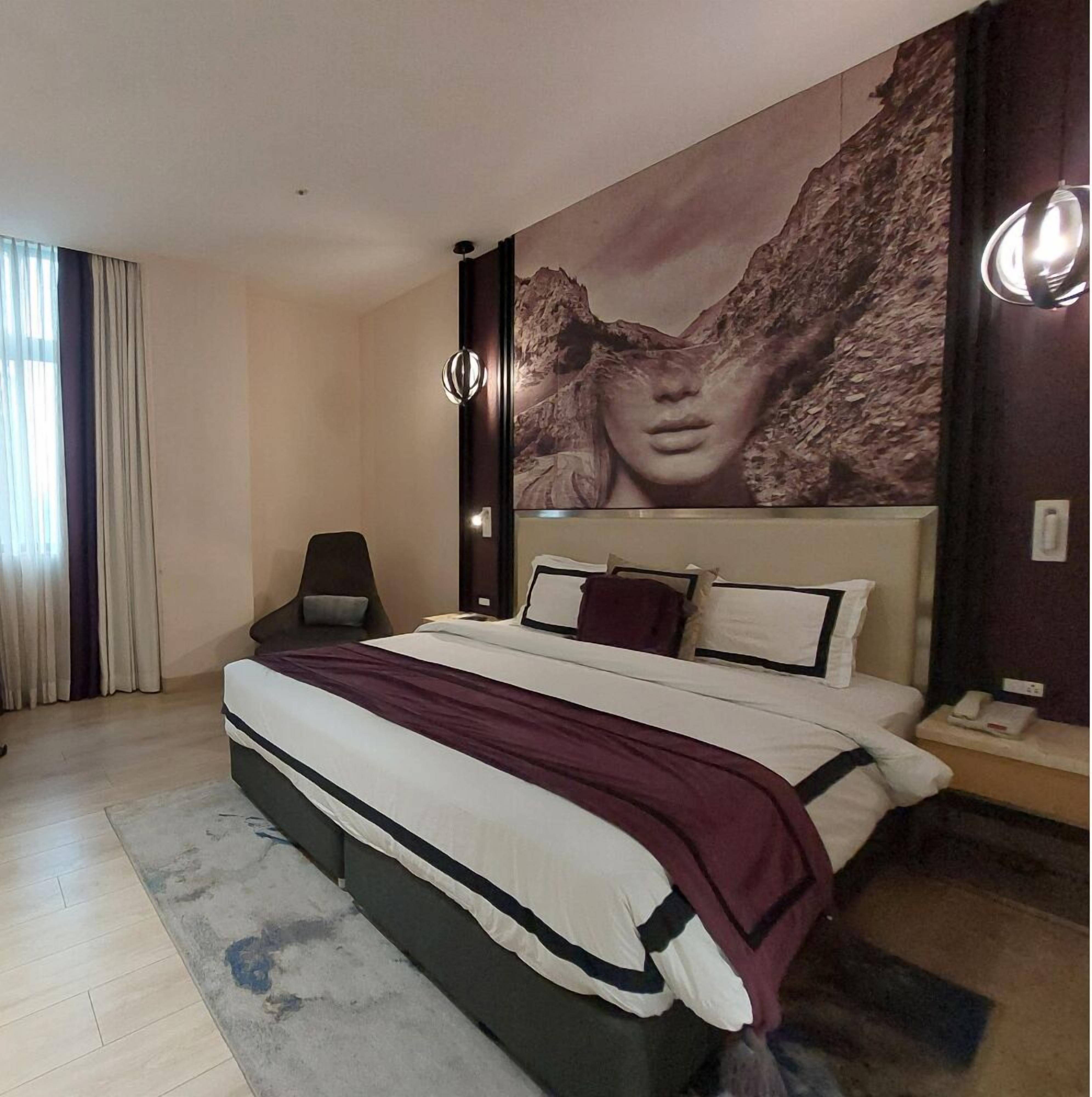 A modern hotel room with a large bed, a nightstand, a chair, and a large wall mural.