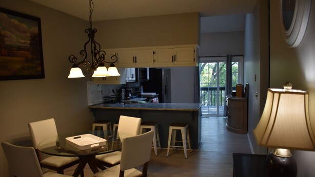 A modern kitchen with white cabinets, a central island, and bar stools. There is a dining table with chairs and a lamp on a side table.
