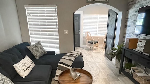 Un soggiorno con un divano nero, un tavolino in legno e un mobile TV con televisore a schermo piatto. Sopra la porta, un cartello con scritto 'Welcome to our beach house'.