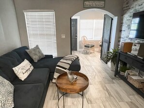 Un soggiorno con un divano nero, un tavolino in legno e un mobile TV con televisore a schermo piatto. Sopra la porta, un cartello con scritto 'Welcome to our beach house'.