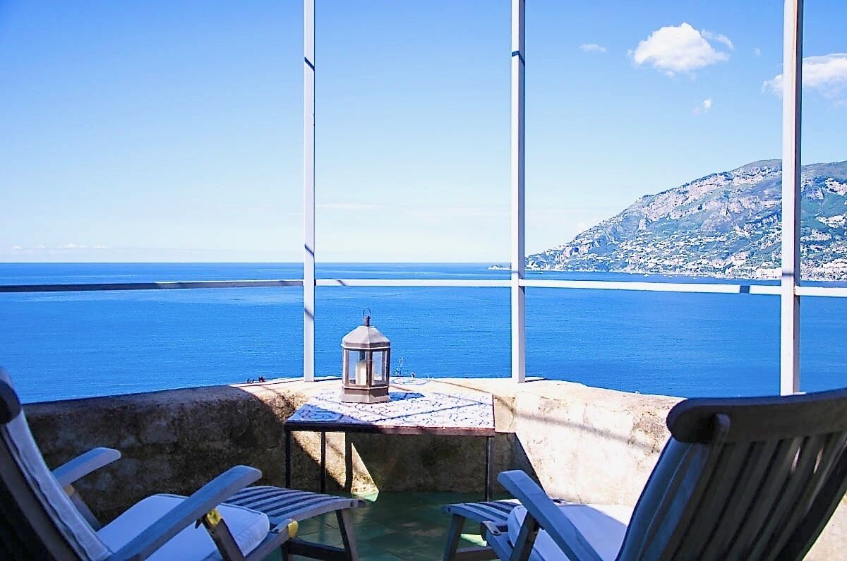 Un balcon avec une table, deux chaises et une lanterne, d’où l’on peut admirer un plan d’eau et une montagne lointaine.