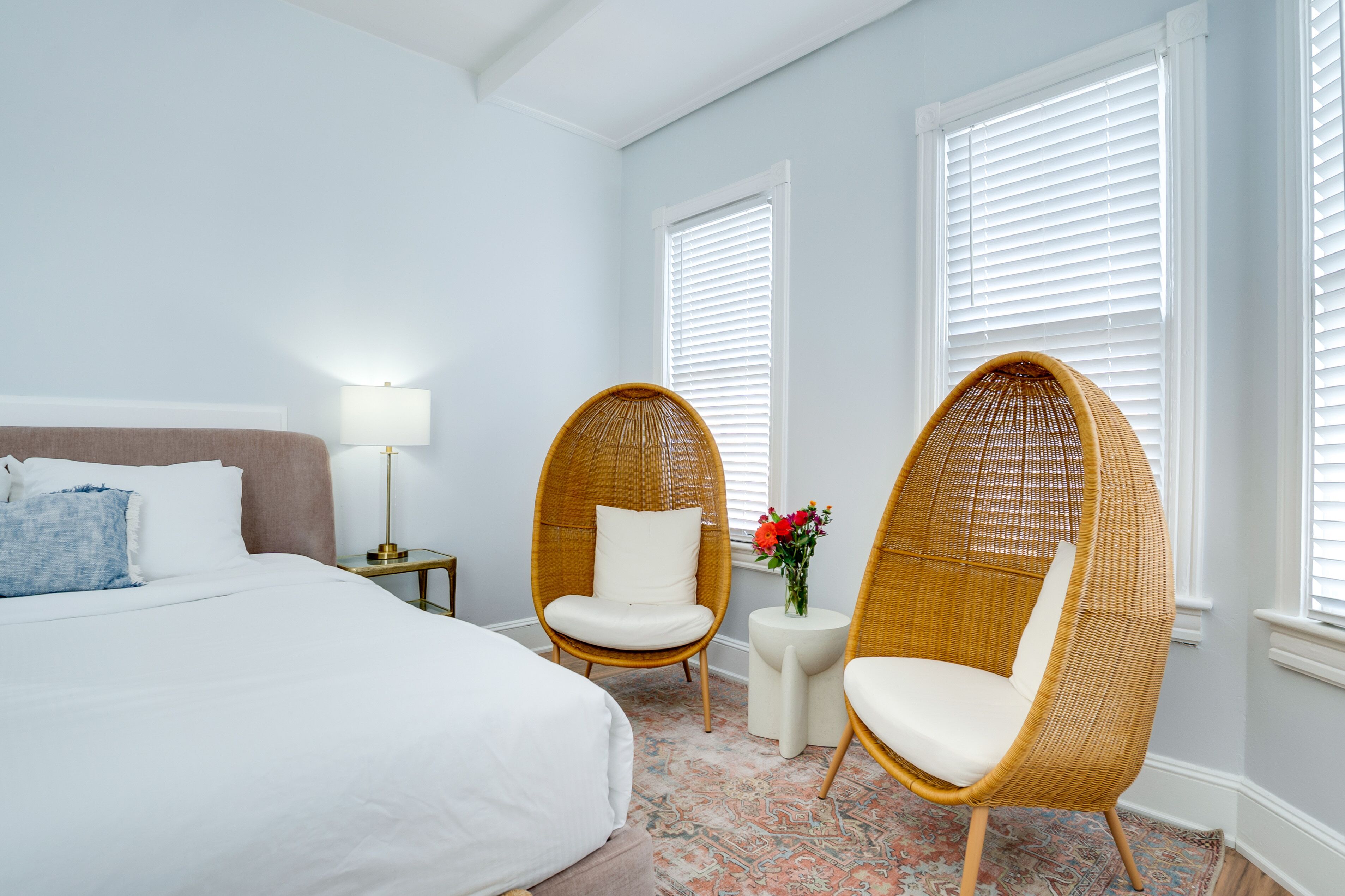 A bedroom with a large bed, two wicker chairs, a small table with a vase of flowers, and a nightstand with a lamp.