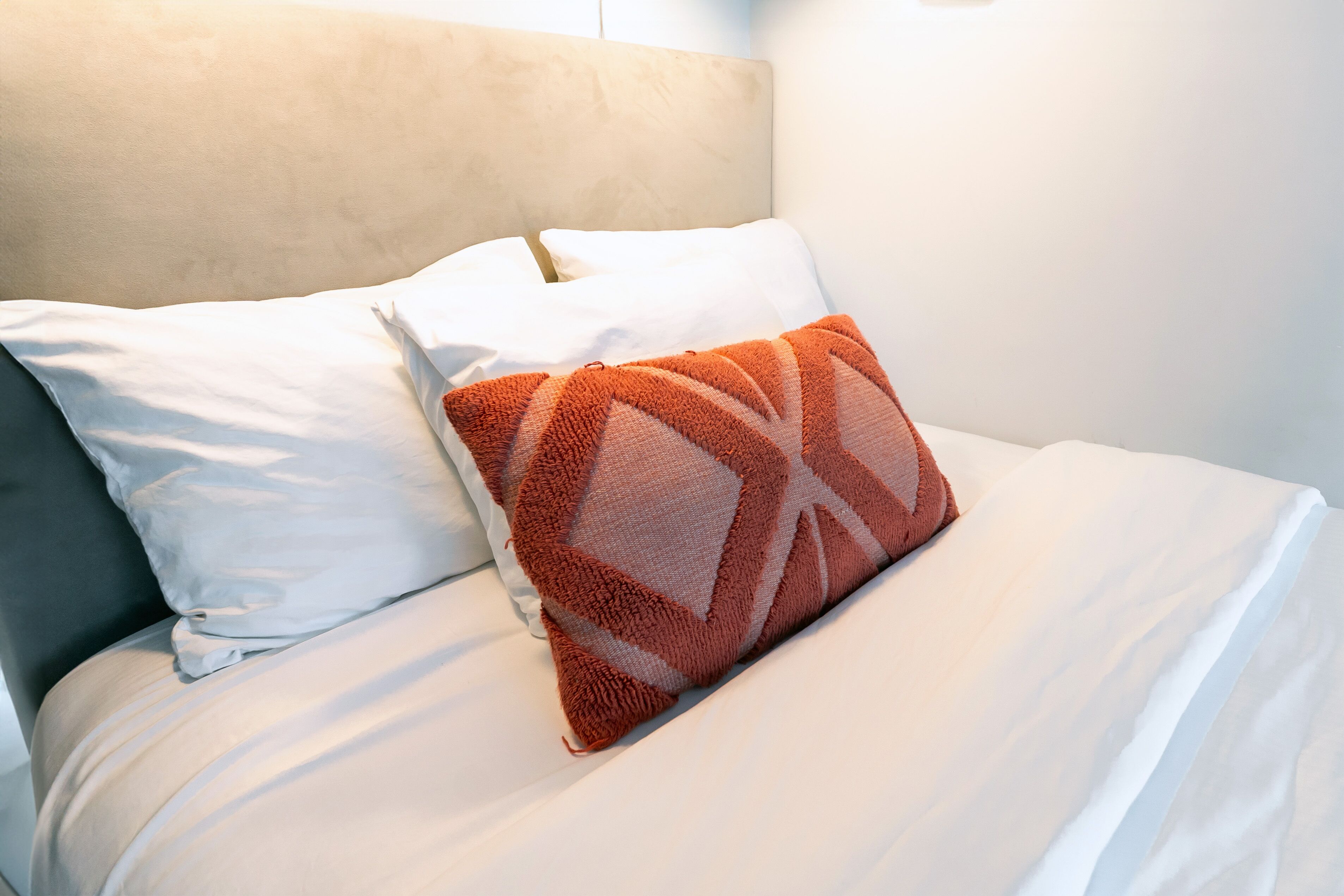A bed with white bedding and a single orange patterned pillow.