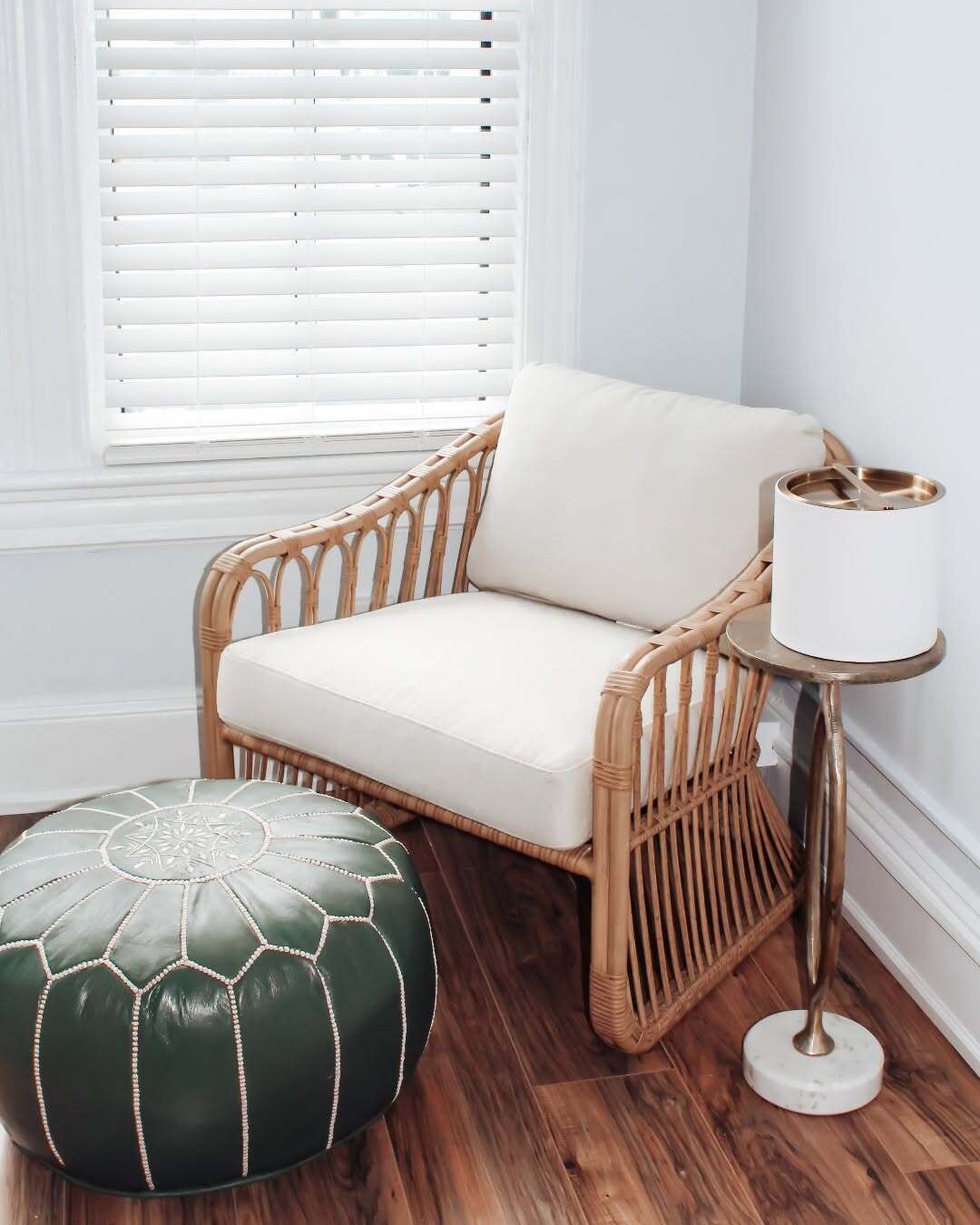 A wicker chair with a cushion, a round ottoman, and a small table with a lamp.