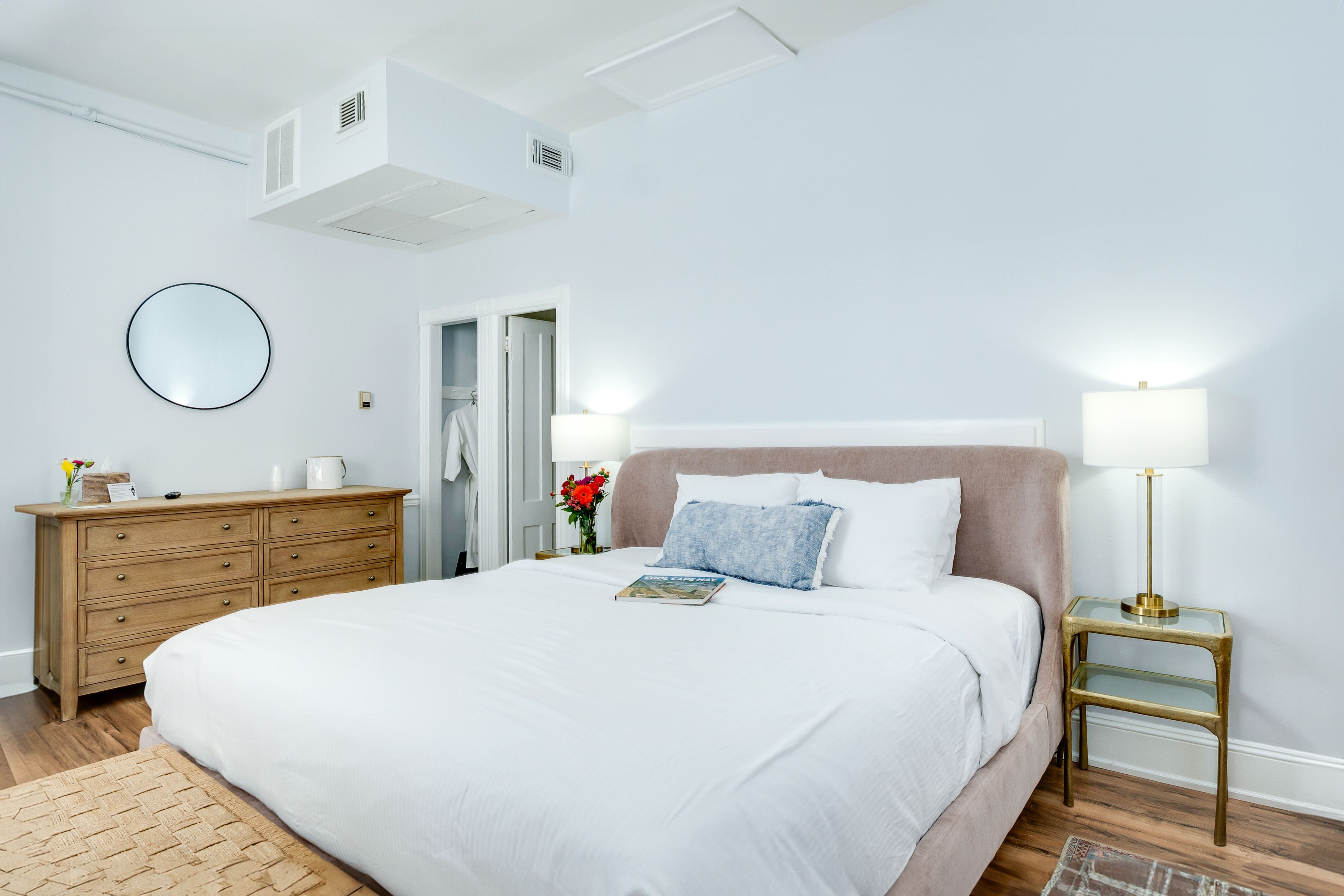 Quarto com uma cama grande, cômoda de madeira, espelho redondo, mesa de cabeceira com abajur e porta que dá para outro ambiente.