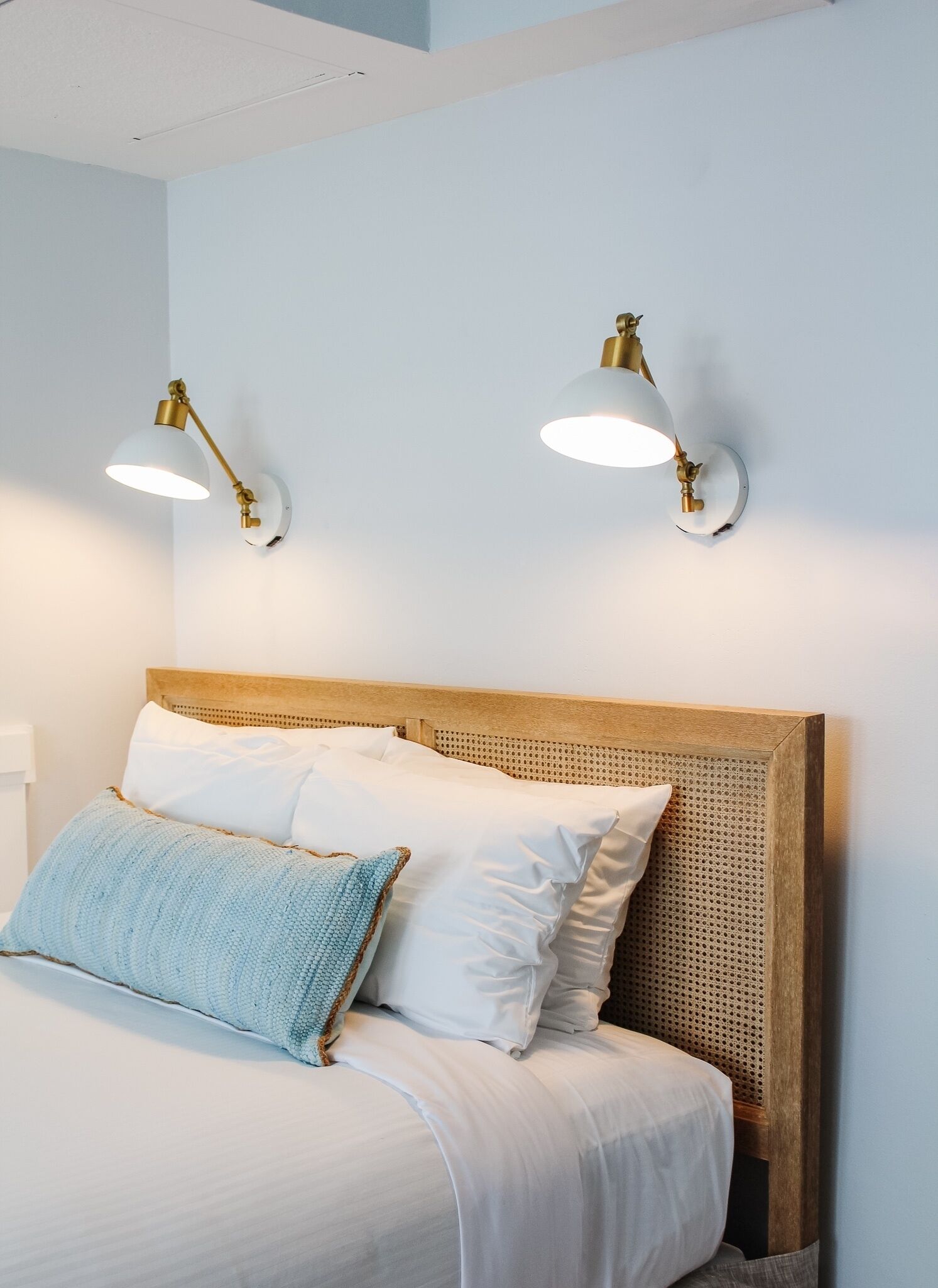 Ein ordentlich bezogenes Bett mit weißen Leinen und einem blauen Akzentkissen, ein Holz-Kopfteil sowie zwei an der Wand befestigte Lampen mit Messingfinish.