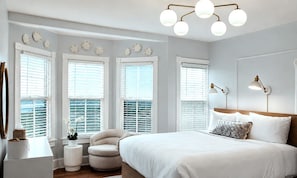 A bedroom with a large bed, bedside lamps, a chair, a small table, and multiple windows with blinds. - The Inn of Cape May (Cape May)