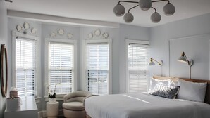 Ein Schlafzimmer mit einem großen Bett, zwei Nachttischlampen, einer Deckenleuchte und drei Fenstern mit Jalousien.