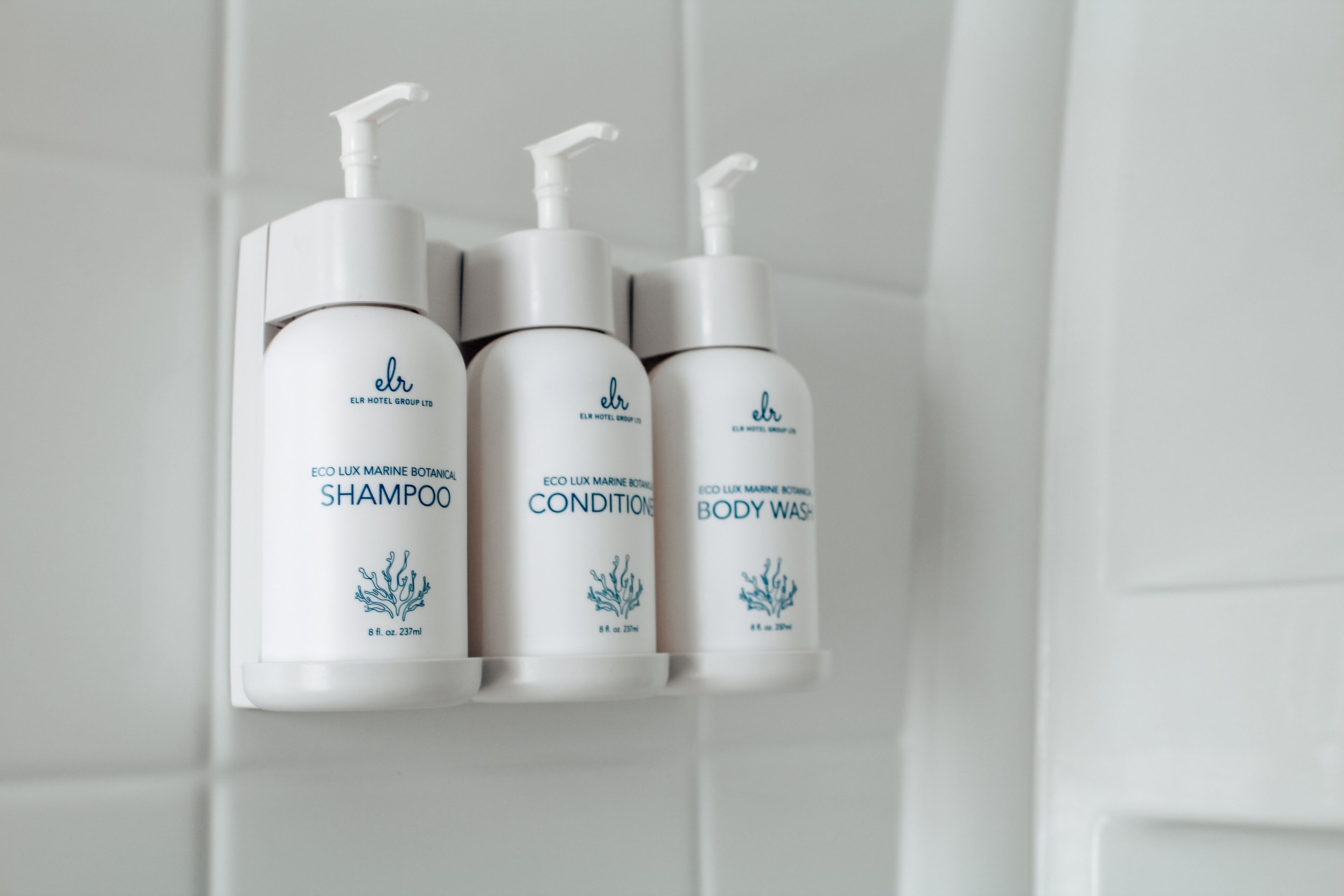 Three bottles of eco-lux marine botanical shampoo, conditioner, and body wash on a white tiled wall.