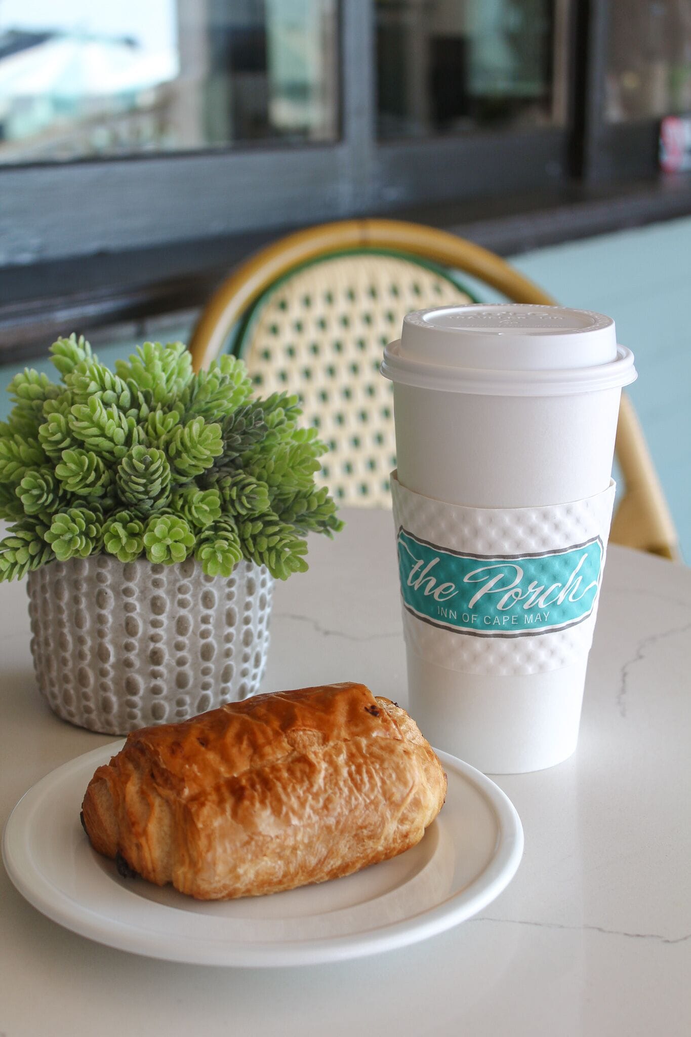 Un croissant sur une assiette, une tasse avec une étiquette et une petite plante en pot sur une table.