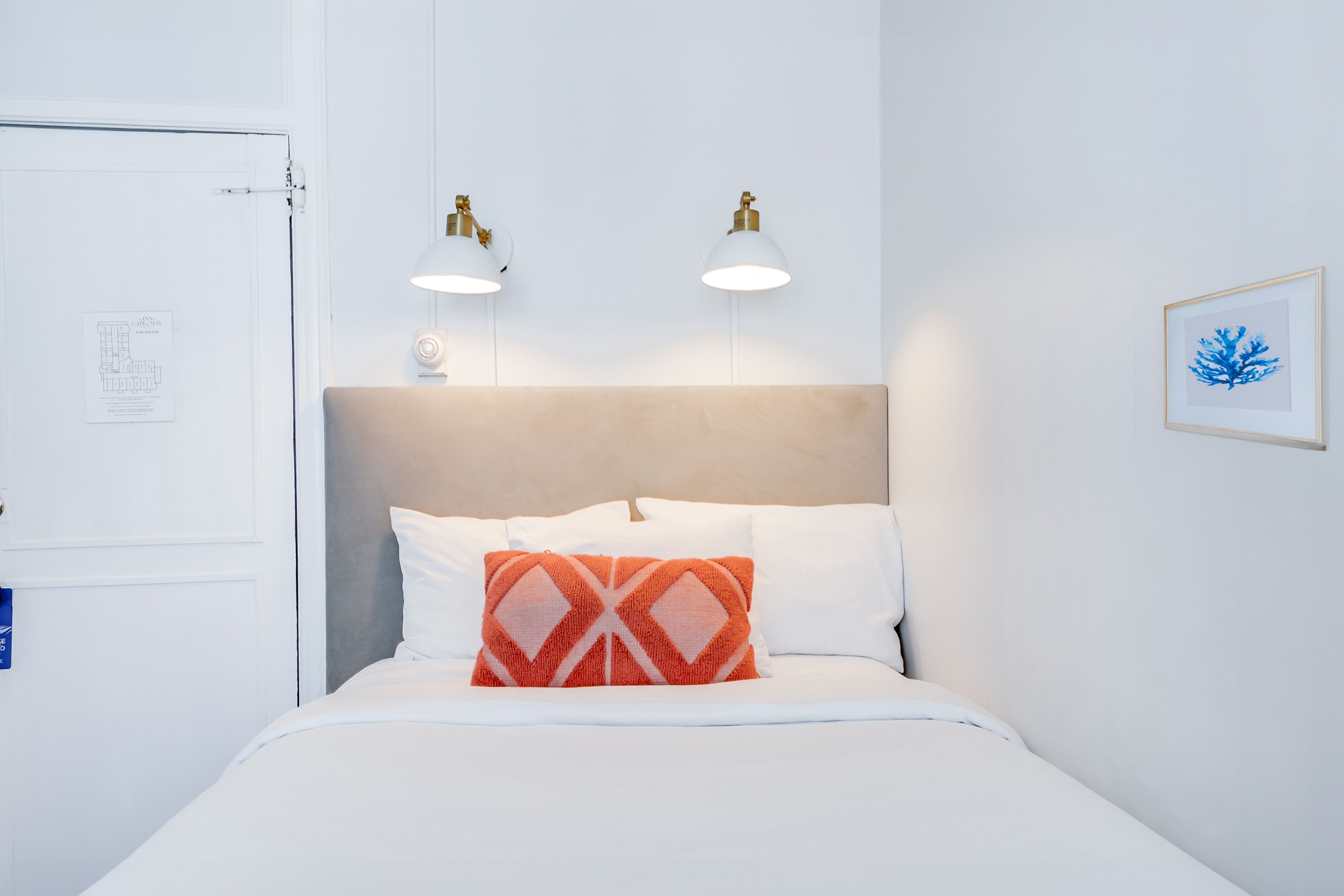 A hotel room with a bed, a headboard, two wall-mounted lamps, a framed picture, and a door.