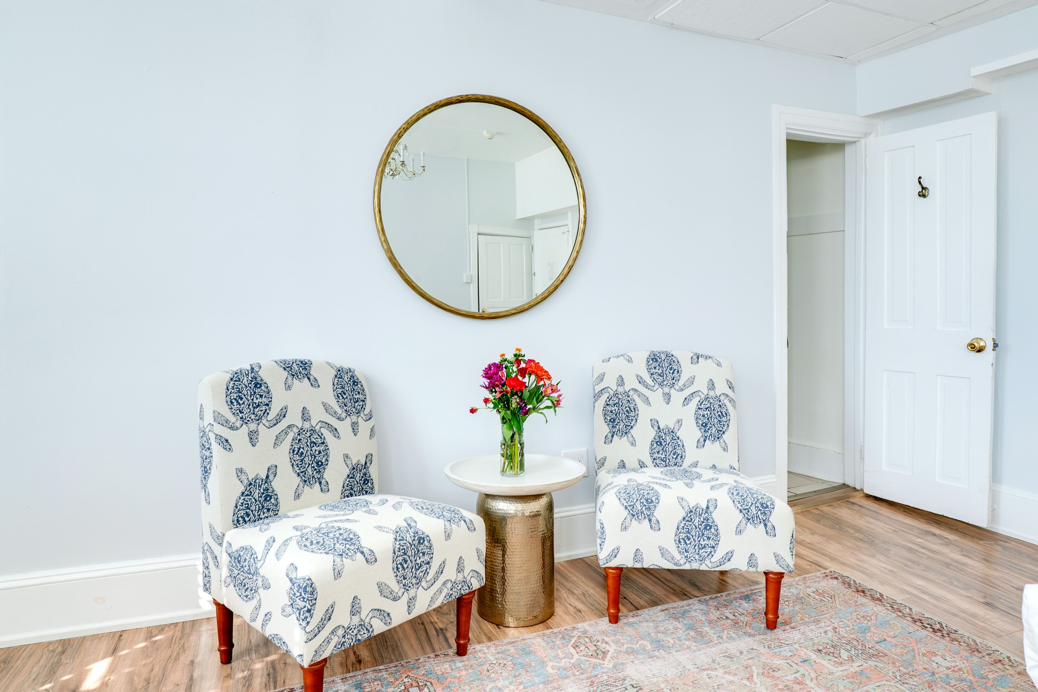 Una habitación con dos sillones estampados, un espejo redondo, una pequeña mesa con un florero y un tapete con diseño.