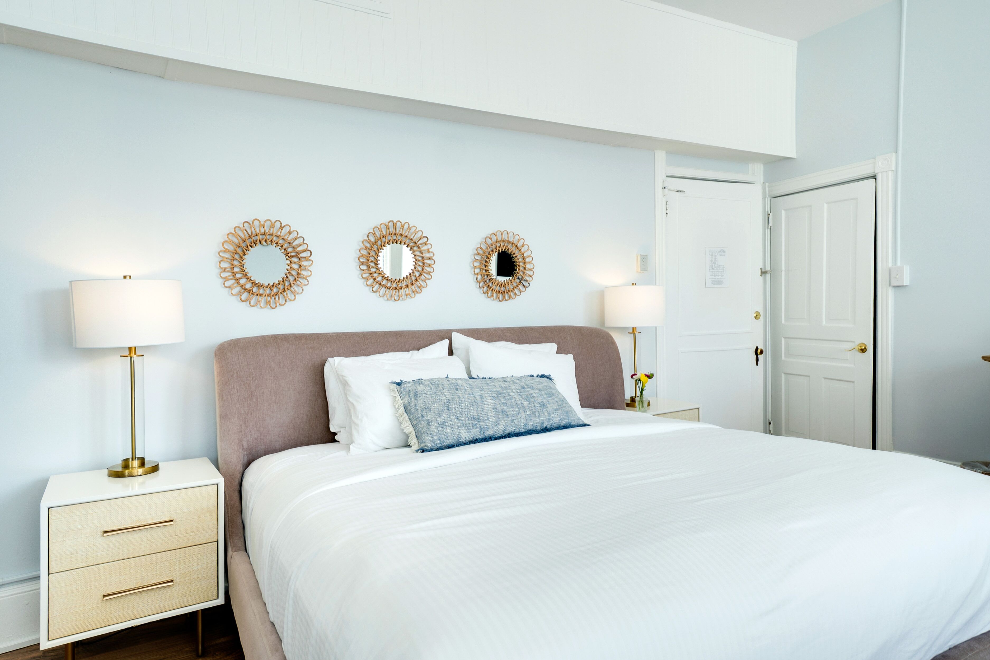 A bedroom with a large bed, two bedside tables with lamps, a door, and three wall-mounted mirrors.