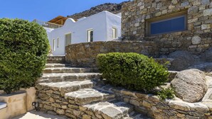 A stone staircase leading to a white building with a wooden roof, surrounded by greenery and a clear blue sky. - Beautiful Villa Isabella (Mykonos)