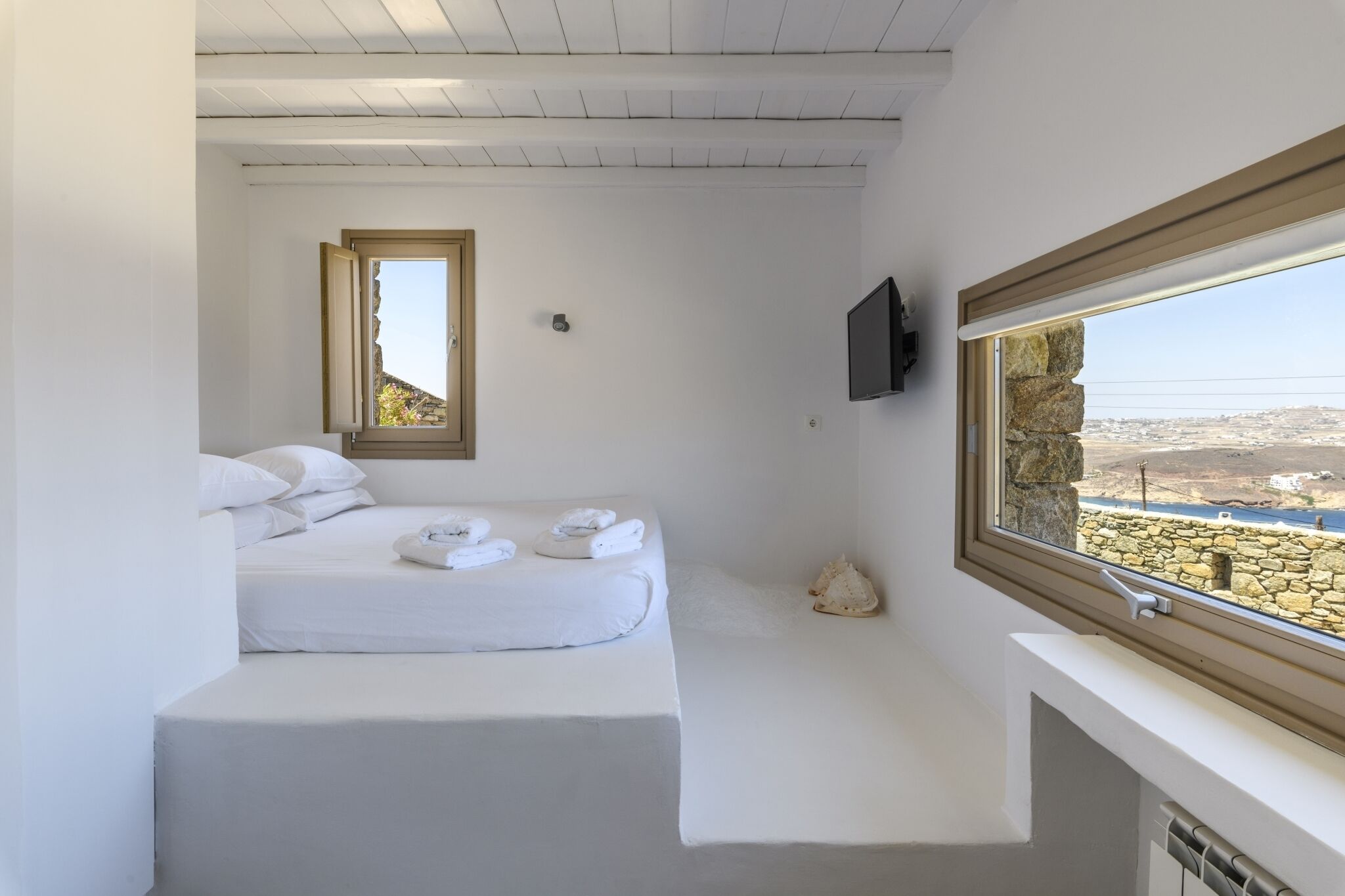 Ein schlicht eingerichtetes Schlafzimmer mit Bett, Fenster und Blick auf eine Steinsäule.