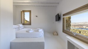 A minimalist bedroom with a bed, a window with a stone wall view, and a mounted TV. - Beautiful Villa Isabella (Mykonos)