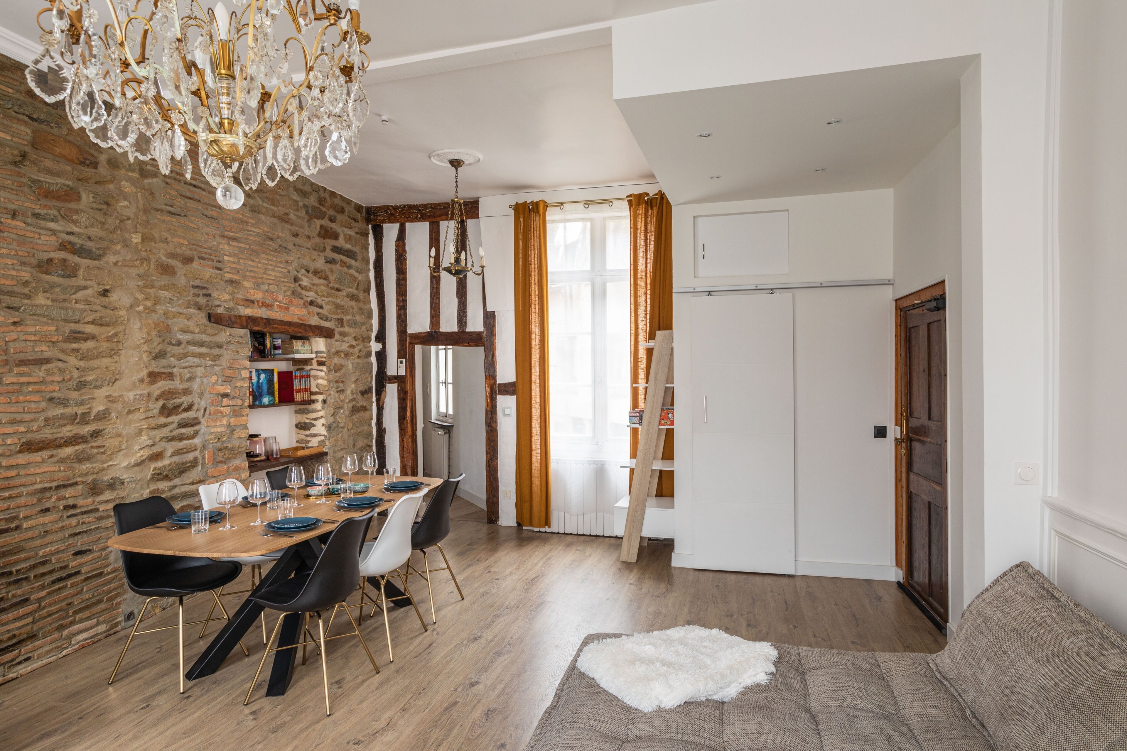 Un coin repas avec une table en bois, des chaises et un lustre. On y trouve un mur en pierre, une fenêtre avec des rideaux et une porte qui donne sur une autre pièce.