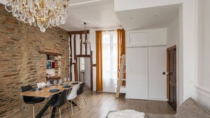 Un comedor con una mesa de madera, sillas y una araña. Se observa una pared de piedra, una ventana con cortinas y una puerta que da a otra habitación.