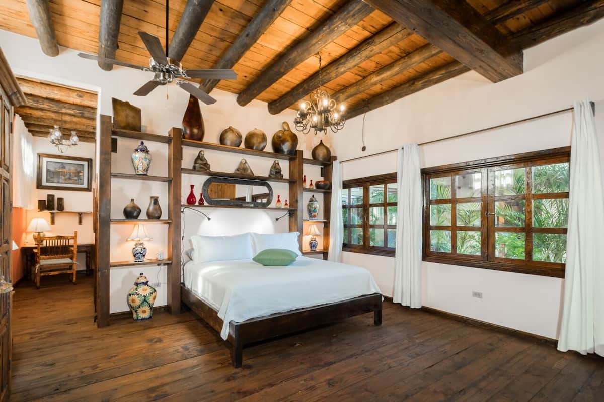 A wooden-floored bedroom with a bed, a chandelier, and a large window with curtains.