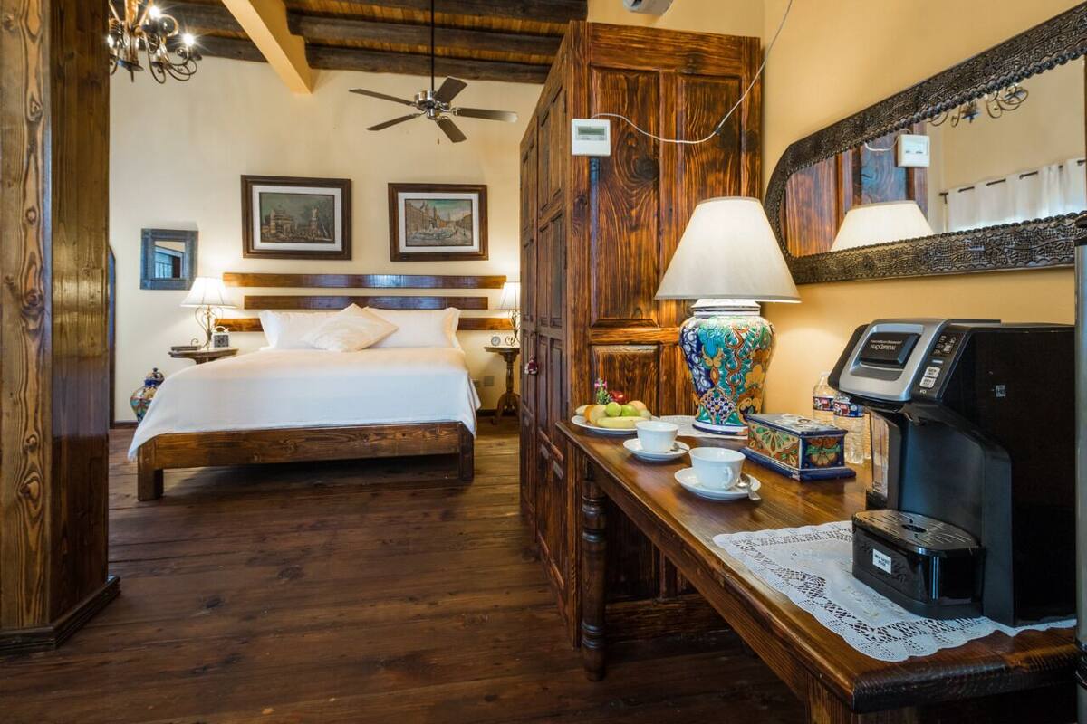 A hotel room with a wooden bed, a desk with a coffee machine, a lamp, and a mirror.