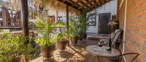 A covered outdoor patio with wooden flooring, potted plants, a round table with chairs, and a bottle of wine.