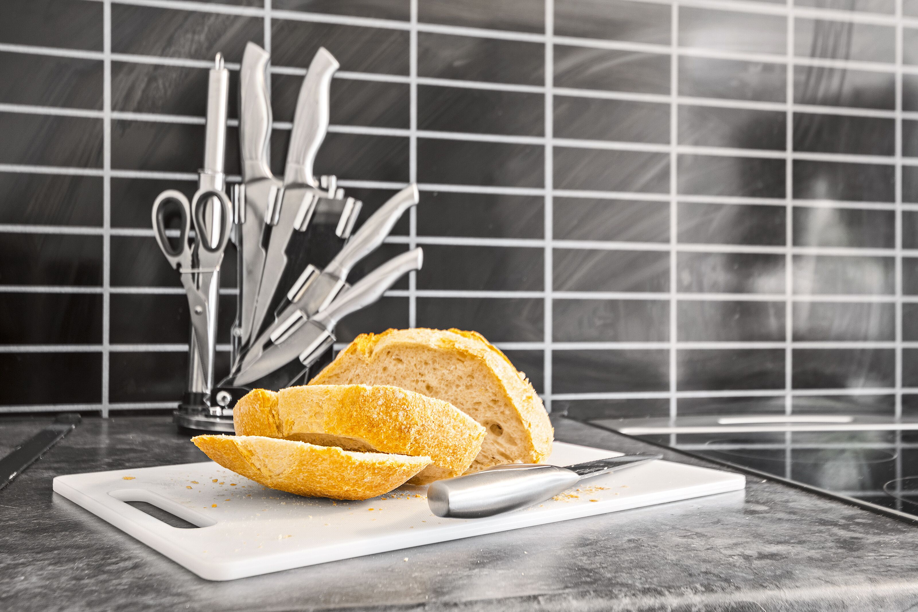 A loaf of bread on a white cutting board with a knife and scissors, set against a tiled backsplash.