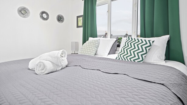 Un lit bien fait, avec une couverture grise, des rideaux verts et des coussins à motifs, dans une chambre moderne dotée d’une grande fenêtre.
