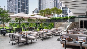 An outdoor dining area with tables and chairs, surrounded by greenery and modern buildings.