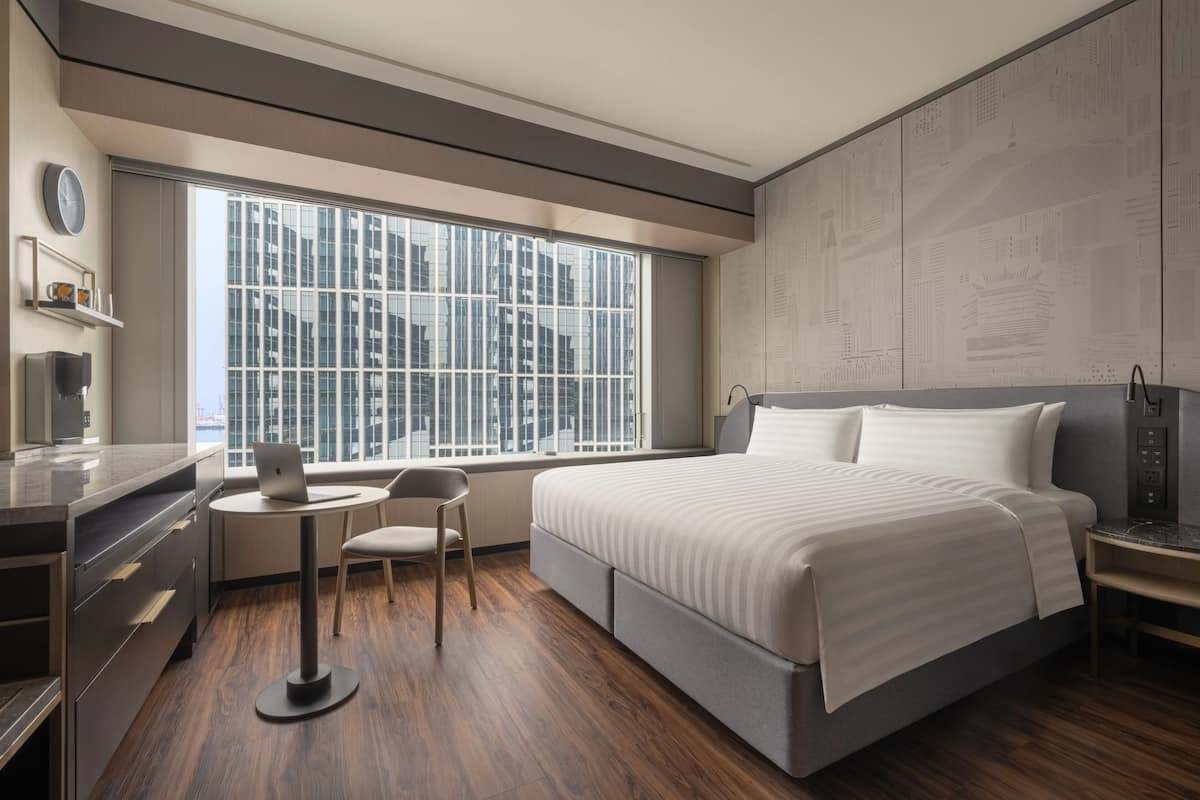 A modern hotel room with a large bed, a desk, a chair, and a window with a city view.