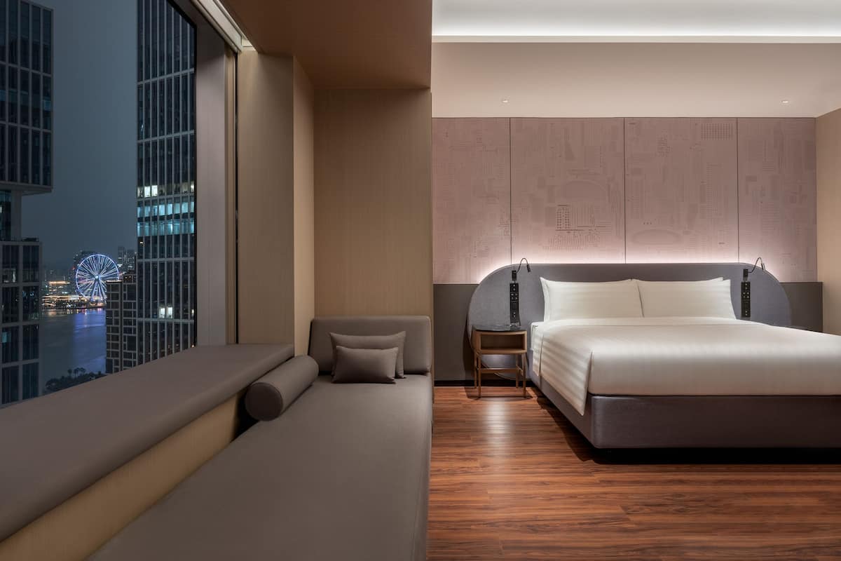 A modern hotel room with a large bed, a sofa, a nightstand, and a view of the cityscape.