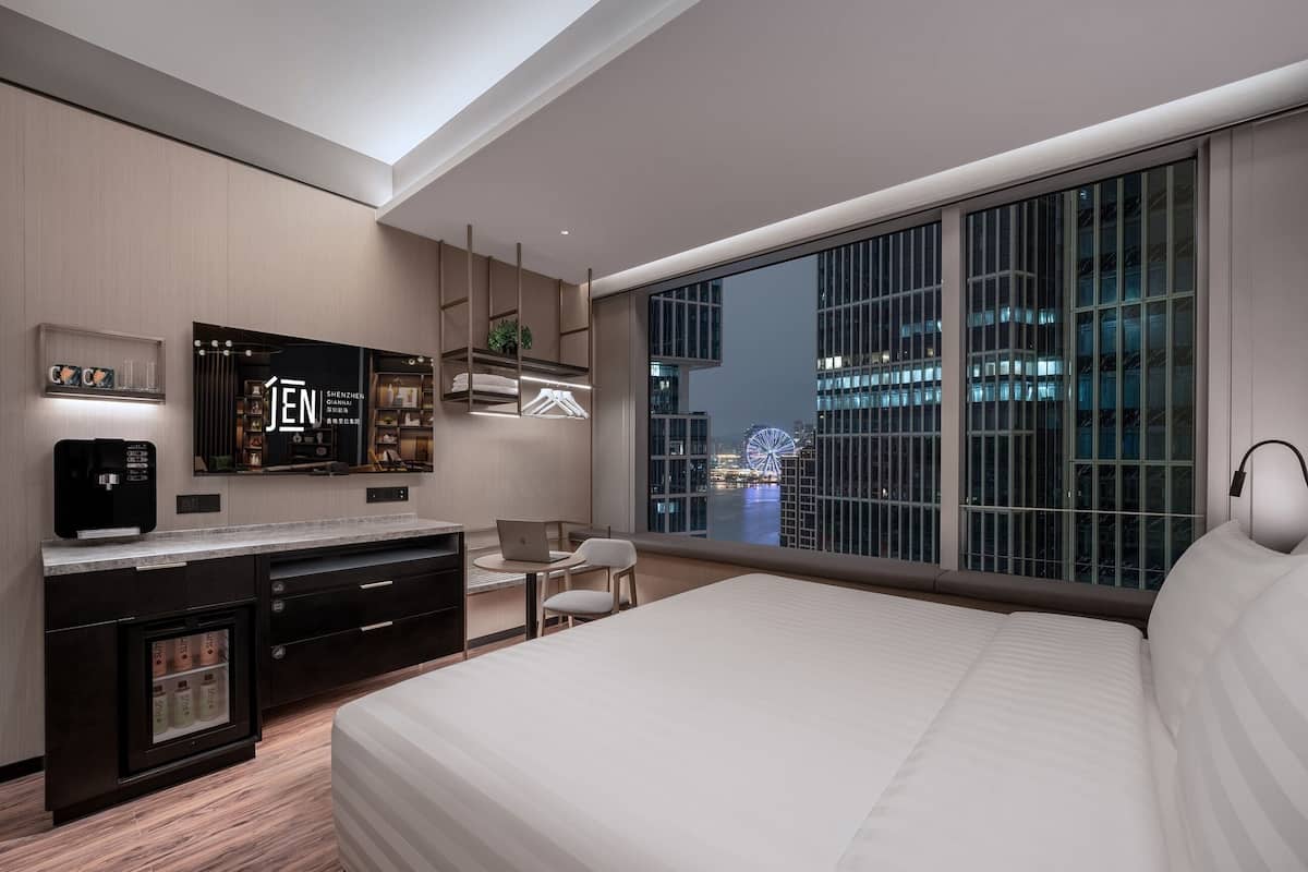 A modern hotel room with a large bed, a desk with a laptop, a minibar, and a view of a cityscape at night.