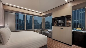 A modern hotel room with a large bed, a seating area, a kitchenette, and a city view.