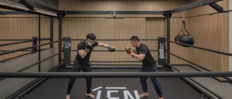 A boxing ring with "JEN" branding, surrounded by black metal railings, in a room with wooden paneling and a hanging punching bag.