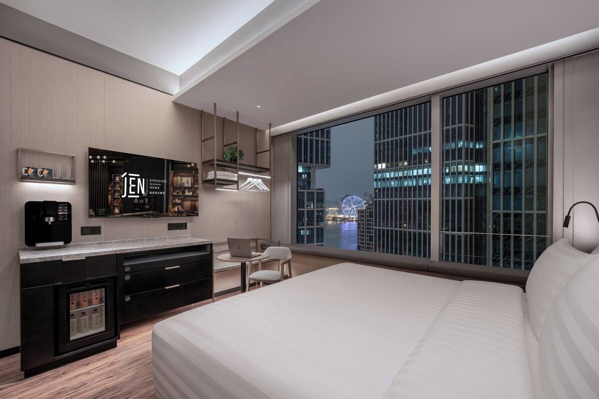 A modern hotel room with a large bed, a desk with a laptop, a minibar, and a view of a cityscape and Ferris wheel at night.