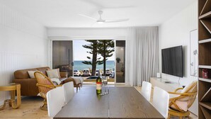A modern living room with a wooden dining table, white chairs, a brown sofa, and a TV on the wall. - Luxury Beachfront Retreat | Henley Beach Esplanade (Henley Beach)