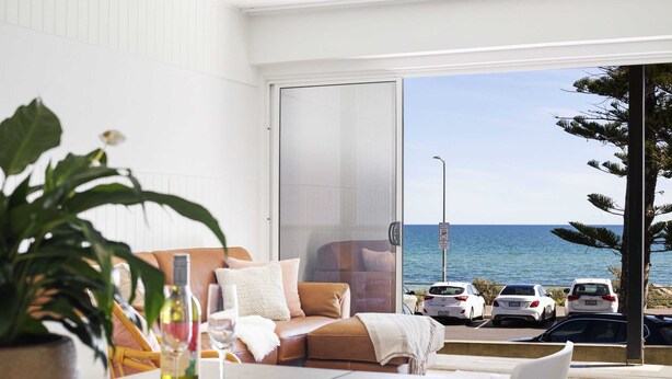 A modern living room with a sofa, a dining table, and a view of the sea and parked cars. - Luxury Beachfront Retreat | Henley Beach Esplanade (Henley Beach)