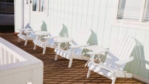 Un hilera de sillas Adirondack blancas sobre una terraza de madera, con un edificio blanco al fondo.