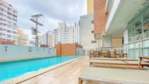 An outdoor swimming pool with lounge chairs and a glass railing, surrounded by high-rise buildings and construction cranes.