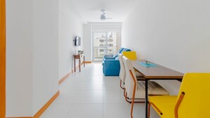 A modern, bright room with a dining table, yellow chairs, a blue sofa, and a TV on the wall.