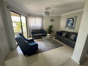 A living room with a sofa, armchair, and coffee table, a ceiling fan, and a large window with curtains.