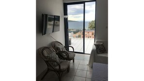 A balcony with a view of buildings and mountains, two wicker chairs with cushions, and a TV mounted on the wall. - Sea View flat - Marmari village 1hour from Rafina (marmari)