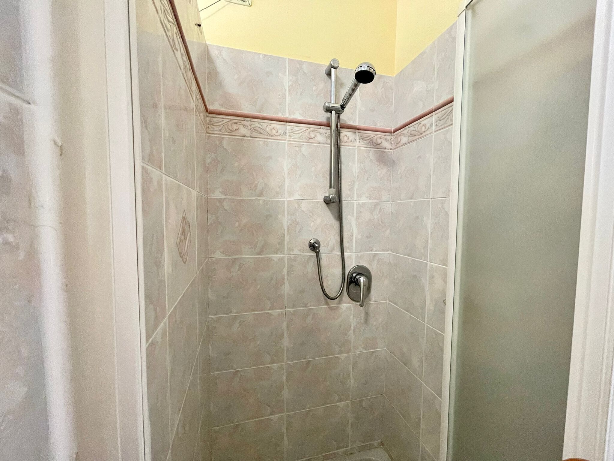 A shower with a handheld showerhead and a wall-mounted faucet in a corner of a room with tiled walls.