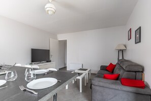 Uma sala de estar moderna com um sofá cinzento, uma mesa de jantar posta com pratos e copos, uma televisão e um abajur.